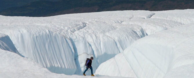 a person walks near an ice canyon on a glacier with mountains in the background